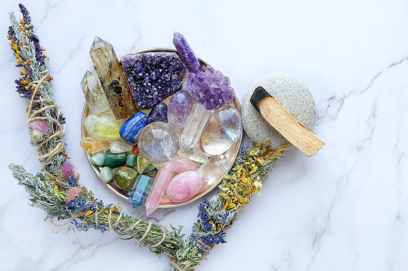 bowl of different multi coloured crystals. bundles og herbs and flowers under and slightly to either side 